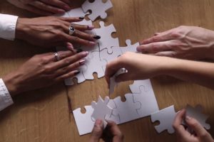 Above close up view hands multi-ethnic business people teammates connects together white pieces of jigsaw puzzle on office desk, participating team building activity, search solutions, synergy concept